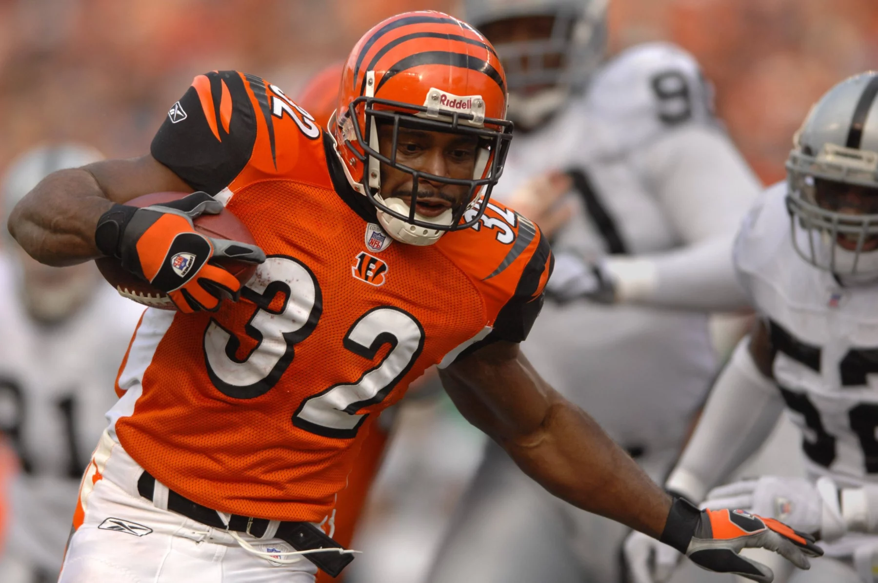 Ein Footballspieler der Cincinnati Bengals, Rudi Johnson, trägt ein orangefarbenes Trikot mit der Nummer 32. Er läuft mit dem Ball und weicht Verteidigern in Weiß und Silber aus. Der Ex-Bengals-Star rennt über das Feld, während im Hintergrund unscharfe Spieler zu sehen sind. Diese Beschreibung wurde mit der FootballR KI automatisch generiert.