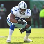 Der Spieler der Dallas Cowboys, Tyler Smith, trägt die Nummer 73 und hockt in einer vorbereiteten Position auf dem grünen Feld. Er trägt einen silbernen Helm, blaue Handschuhe und eine weiße Hose mit blauen Streifen. Im Hintergrund sind unscharfe Spieler und Mitarbeiter an der Seitenlinie zu sehen. Diese Beschreibung wurde mit der FootballR KI automatisch generiert.