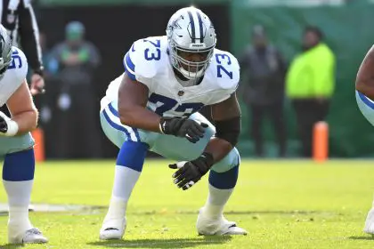 Der Spieler der Dallas Cowboys, Tyler Smith, trägt die Nummer 73 und hockt in einer vorbereiteten Position auf dem grünen Feld. Er trägt einen silbernen Helm, blaue Handschuhe und eine weiße Hose mit blauen Streifen. Im Hintergrund sind unscharfe Spieler und Mitarbeiter an der Seitenlinie zu sehen. Diese Beschreibung wurde mit der FootballR KI automatisch generiert.