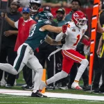 Der Kansas City Chiefs-Spieler Xavier Worthy in einem weiß-roten Trikot läuft mit dem Football, während ein Spieler der Philadelphia Eagles in Grün versucht, ihn zu stoppen. Das Personal an der Seitenlinie und die Schiedsrichter beobachten das Geschehen auf dem Footballfeld. Diese Beschreibung wurde mit der FootballR KI automatisch generiert.