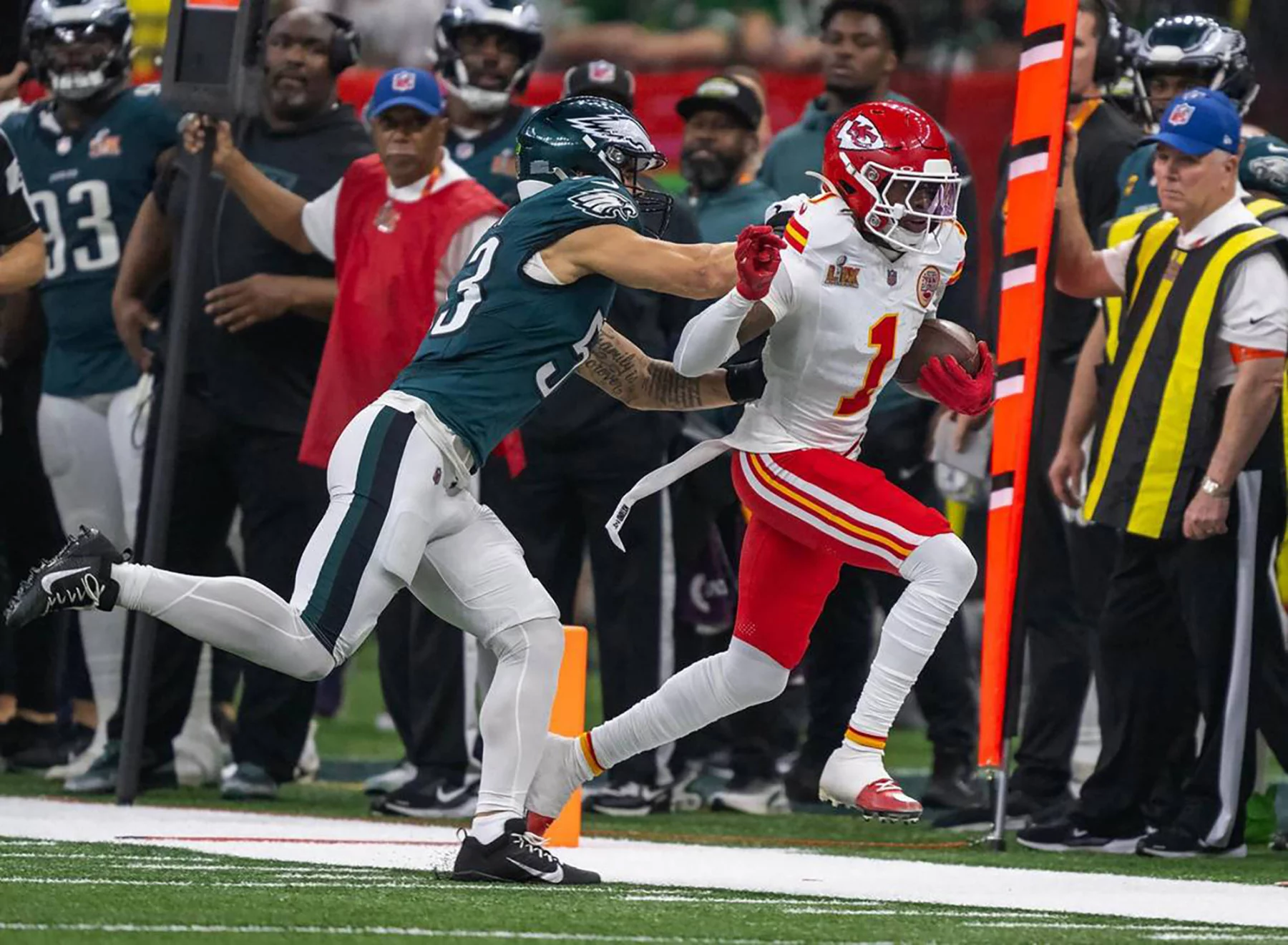 Der Kansas City Chiefs-Spieler Xavier Worthy in einem weiß-roten Trikot läuft mit dem Football, während ein Spieler der Philadelphia Eagles in Grün versucht, ihn zu stoppen. Das Personal an der Seitenlinie und die Schiedsrichter beobachten das Geschehen auf dem Footballfeld. Diese Beschreibung wurde mit der FootballR KI automatisch generiert.