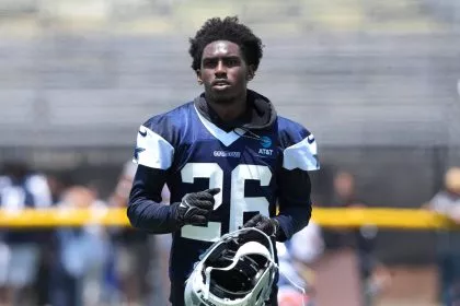 Ein Footballspieler, identifiziert als DaRon Bland, trägt ein marineblaues und weißes Trikot der Dallas Cowboys mit der Nummer 26. Er steht auf dem Spielfeld, hält seinen Helm, hat kurzes lockiges Haar und einen konzentrierten Gesichtsausdruck, mit unscharfen Tribünen und Zuschauern im Hintergrund. Diese Beschreibung wurde mit der FootballR KI automatisch generiert.