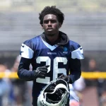 Ein Footballspieler, identifiziert als DaRon Bland, trägt ein marineblaues und weißes Trikot der Dallas Cowboys mit der Nummer 26. Er steht auf dem Spielfeld, hält seinen Helm, hat kurzes lockiges Haar und einen konzentrierten Gesichtsausdruck, mit unscharfen Tribünen und Zuschauern im Hintergrund. Diese Beschreibung wurde mit der FootballR KI automatisch generiert.