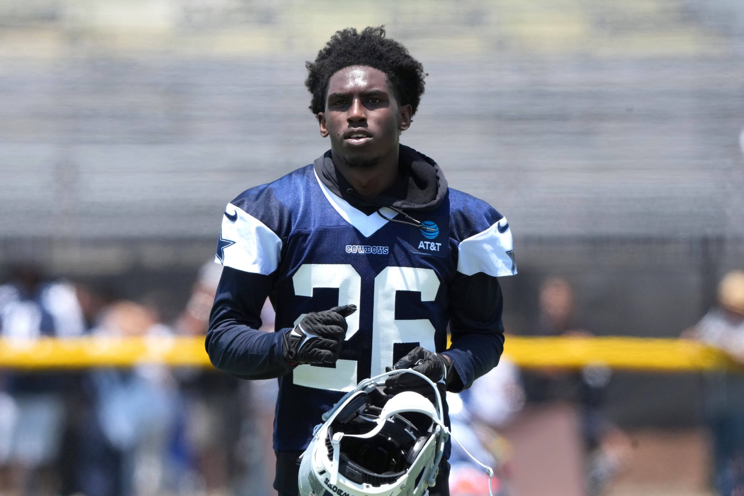 Ein Footballspieler, identifiziert als DaRon Bland, trägt ein marineblaues und weißes Trikot der Dallas Cowboys mit der Nummer 26. Er steht auf dem Spielfeld, hält seinen Helm, hat kurzes lockiges Haar und einen konzentrierten Gesichtsausdruck, mit unscharfen Tribünen und Zuschauern im Hintergrund. Diese Beschreibung wurde mit der FootballR KI automatisch generiert.