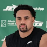 Alijah Vera-Tucker mit kurzen lockigen Haaren, einem Spitzbart und einem ernsten Gesichtsausdruck sitzt vor einem grünen Hintergrund mit "Jets"- und Sponsorenlogos. Er trägt ein ärmelloses schwarzes Sporthemd mit einem weißen Jets-Logo auf der Brust. Diese Beschreibung wurde mit der FootballR KI automatisch generiert.