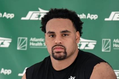 Alijah Vera-Tucker mit kurzen lockigen Haaren, einem Spitzbart und einem ernsten Gesichtsausdruck sitzt vor einem grünen Hintergrund mit "Jets"- und Sponsorenlogos. Er trägt ein ärmelloses schwarzes Sporthemd mit einem weißen Jets-Logo auf der Brust. Diese Beschreibung wurde mit der FootballR KI automatisch generiert.