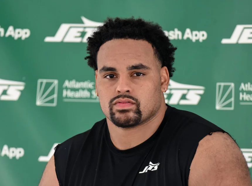 Alijah Vera-Tucker mit kurzen lockigen Haaren, einem Spitzbart und einem ernsten Gesichtsausdruck sitzt vor einem grünen Hintergrund mit "Jets"- und Sponsorenlogos. Er trägt ein ärmelloses schwarzes Sporthemd mit einem weißen Jets-Logo auf der Brust. Diese Beschreibung wurde mit der FootballR KI automatisch generiert.