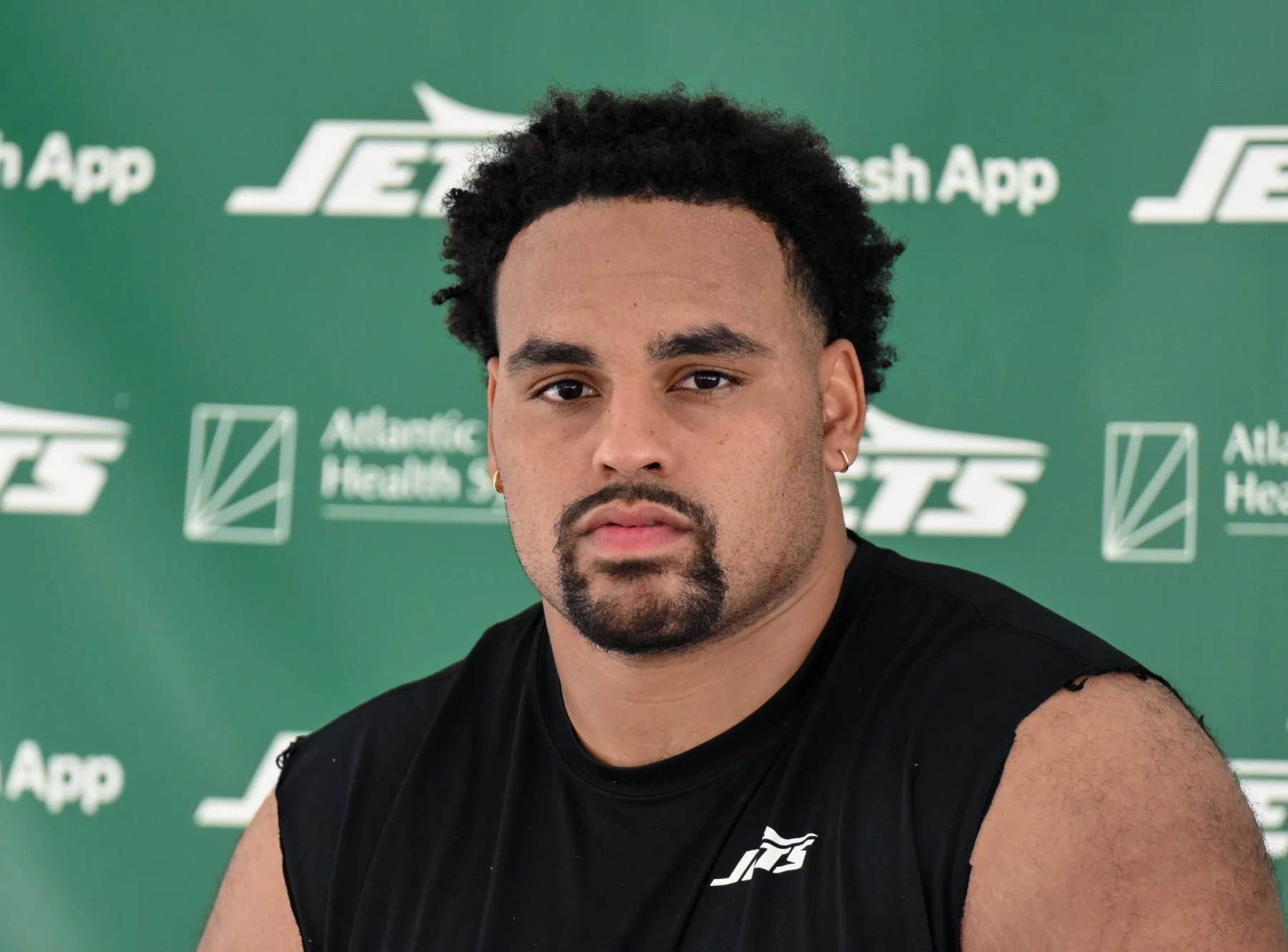 Alijah Vera-Tucker mit kurzen lockigen Haaren, einem Spitzbart und einem ernsten Gesichtsausdruck sitzt vor einem grünen Hintergrund mit "Jets"- und Sponsorenlogos. Er trägt ein ärmelloses schwarzes Sporthemd mit einem weißen Jets-Logo auf der Brust. Diese Beschreibung wurde mit der FootballR KI automatisch generiert.