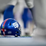 Shane Bowen - Trade-Deadline - Ein blauer Football-Helm der New York Giants mit der Nummer 6 liegt auf dem Spielfeld, umgeben von unscharfen Spielerbeinen. Der Helm trägt das ikonische Teamlogo, einen roten Streifen und Aufkleber und zeigt auf der Rückseite das NFL-Schild und die amerikanische Flagge. Diese Beschreibung wurde mit der FootballR KI automatisch generiert.