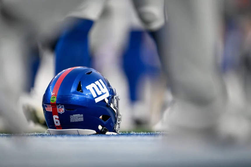 Shane Bowen - Trade-Deadline - Ein blauer Football-Helm der New York Giants mit der Nummer 6 liegt auf dem Spielfeld, umgeben von unscharfen Spielerbeinen. Der Helm trägt das ikonische Teamlogo, einen roten Streifen und Aufkleber und zeigt auf der Rückseite das NFL-Schild und die amerikanische Flagge. Diese Beschreibung wurde mit der FootballR KI automatisch generiert.