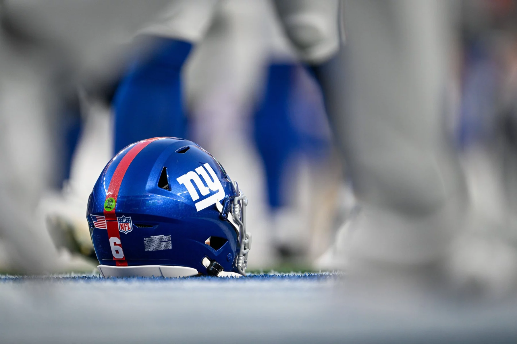 Shane Bowen - Trade-Deadline - Ein blauer Football-Helm der New York Giants mit der Nummer 6 liegt auf dem Spielfeld, umgeben von unscharfen Spielerbeinen. Der Helm trägt das ikonische Teamlogo, einen roten Streifen und Aufkleber und zeigt auf der Rückseite das NFL-Schild und die amerikanische Flagge. Diese Beschreibung wurde mit der FootballR KI automatisch generiert.