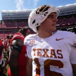 Arch Manning der College Football QB der Texas Longhorns mit weißem Helm und der Trikotnummer 16 steht während der College-Football-Woche 1 zusammen mit anderen Spielern, einem Kameramann in Grün und einem Stadion voller Fans in Rot unter einem sonnigen Himmel auf dem Spielfeld. Diese Beschreibung wurde mit der FootballR KI automatisch generiert.