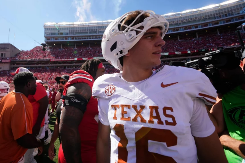 Arch Manning der College Football QB der Texas Longhorns mit weißem Helm und der Trikotnummer 16 steht während der College-Football-Woche 1 zusammen mit anderen Spielern, einem Kameramann in Grün und einem Stadion voller Fans in Rot unter einem sonnigen Himmel auf dem Spielfeld. Diese Beschreibung wurde mit der FootballR KI automatisch generiert.