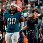 Der Footballspieler der Philadelphia Eagles, Jalen Carter, trägt die Nummer 98 und steht während eines Spiels an der Seitenlinie neben seinen Mannschaftskameraden und Trainern. Er trägt seine Uniform und hat die Hände hinter dem Rücken verschränkt, während im Hintergrund das überfüllte Stadion summt. Diese Beschreibung wurde mit der FootballR KI automatisch generiert.