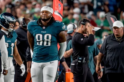 Der Footballspieler der Philadelphia Eagles, Jalen Carter, trägt die Nummer 98 und steht während eines Spiels an der Seitenlinie neben seinen Mannschaftskameraden und Trainern. Er trägt seine Uniform und hat die Hände hinter dem Rücken verschränkt, während im Hintergrund das überfüllte Stadion summt. Diese Beschreibung wurde mit der FootballR KI automatisch generiert.