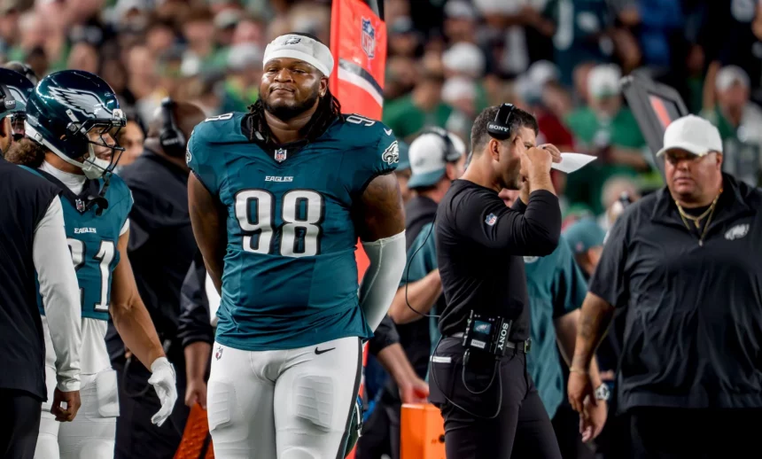 Der Footballspieler der Philadelphia Eagles, Jalen Carter, trägt die Nummer 98 und steht während eines Spiels an der Seitenlinie neben seinen Mannschaftskameraden und Trainern. Er trägt seine Uniform und hat die Hände hinter dem Rücken verschränkt, während im Hintergrund das überfüllte Stadion summt. Diese Beschreibung wurde mit der FootballR KI automatisch generiert.