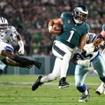 Eagles ringen Cowboys nieder - Der Quarterback Jalen Hurts der Philadelphia Eagles im dunkelgrünen Trikot mit der Nummer 1 läuft mit dem Football und weicht zwei Cowboys-Verteidigern während eines spannenden Saison-Auftaktduells aus. Das Geschehen spielt sich auf einem grünen Feld mit einer unscharfen Menschenmenge im Hintergrund ab. Diese Beschreibung wurde mit der FootballR KI automatisch generiert.