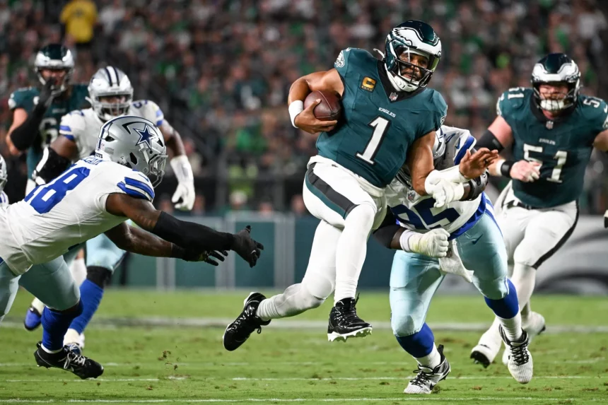 Eagles ringen Cowboys nieder - Der Quarterback Jalen Hurts der Philadelphia Eagles im dunkelgrünen Trikot mit der Nummer 1 läuft mit dem Football und weicht zwei Cowboys-Verteidigern während eines spannenden Saison-Auftaktduells aus. Das Geschehen spielt sich auf einem grünen Feld mit einer unscharfen Menschenmenge im Hintergrund ab. Diese Beschreibung wurde mit der FootballR KI automatisch generiert.