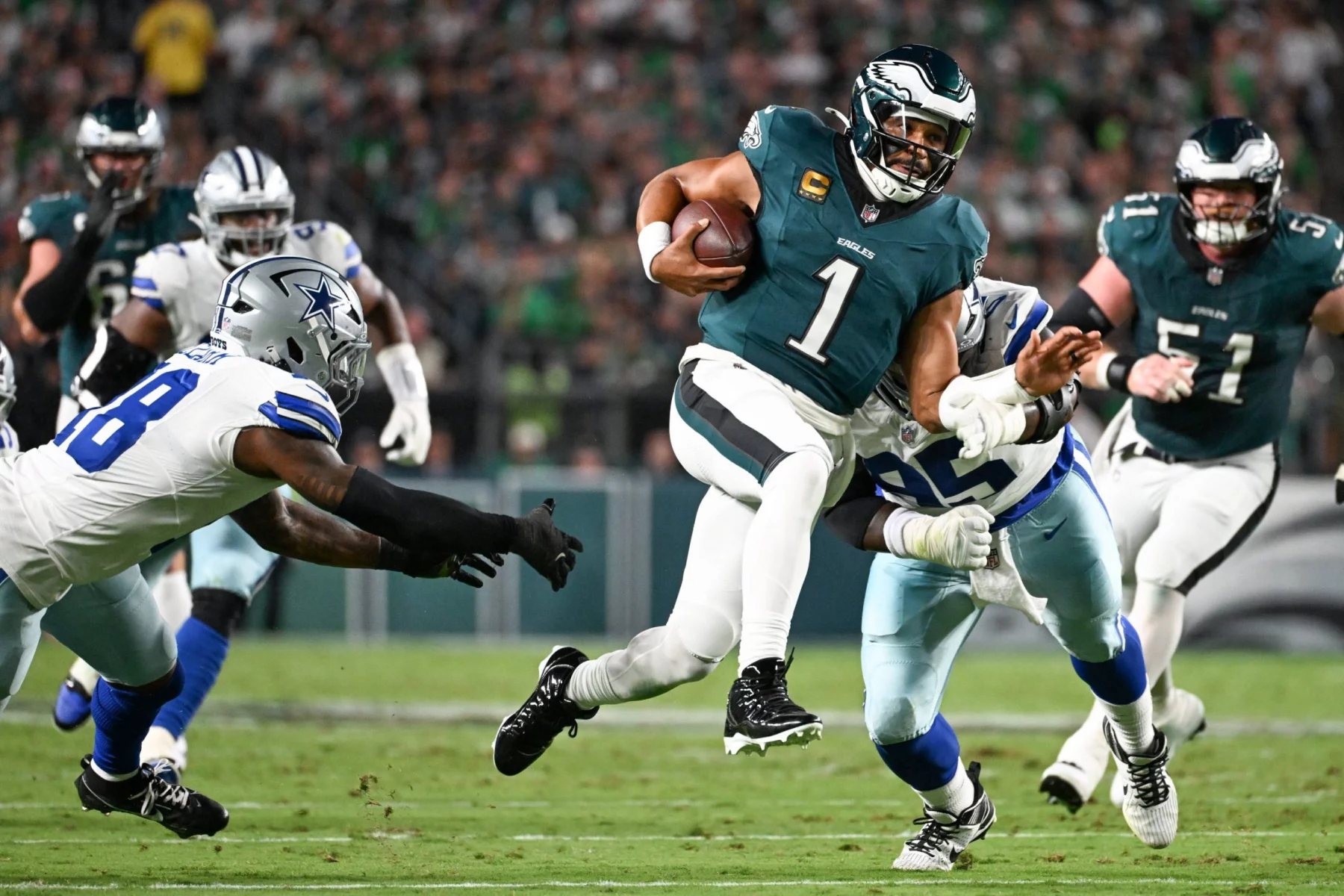 Eagles ringen Cowboys nieder - Der Quarterback Jalen Hurts der Philadelphia Eagles im dunkelgrünen Trikot mit der Nummer 1 läuft mit dem Football und weicht zwei Cowboys-Verteidigern während eines spannenden Saison-Auftaktduells aus. Das Geschehen spielt sich auf einem grünen Feld mit einer unscharfen Menschenmenge im Hintergrund ab. Diese Beschreibung wurde mit der FootballR KI automatisch generiert.