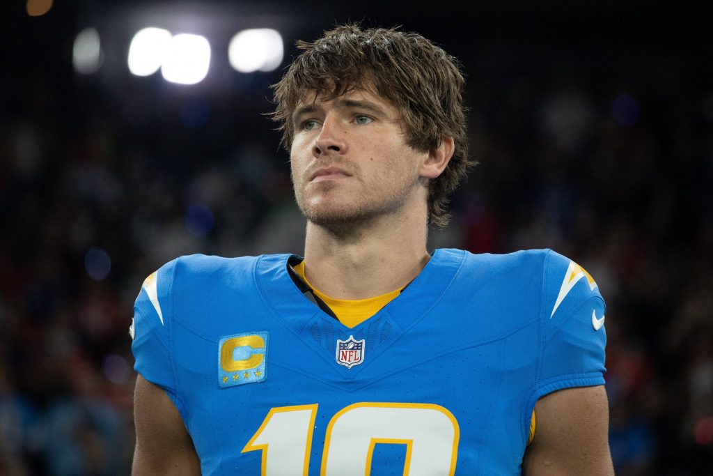 DAZN - Ein Footballspieler, Justin Herbert, trägt ein blaues Los Angeles Chargers-Trikot mit der Nummer 10 und dem Kapitänsaufnäher und steht konzentriert auf dem Spielfeld. Sein braunes Haar ist leicht zerzaust, während die Stadionbeleuchtung und die Zuschauer im Hintergrund verschwimmen. Diese Beschreibung wurde mit der FootballR KI automatisch generiert.