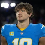 DAZN - Ein Footballspieler, Justin Herbert, trägt ein blaues Los Angeles Chargers-Trikot mit der Nummer 10 und dem Kapitänsaufnäher und steht konzentriert auf dem Spielfeld. Sein braunes Haar ist leicht zerzaust, während die Stadionbeleuchtung und die Zuschauer im Hintergrund verschwimmen. Diese Beschreibung wurde mit der FootballR KI automatisch generiert.