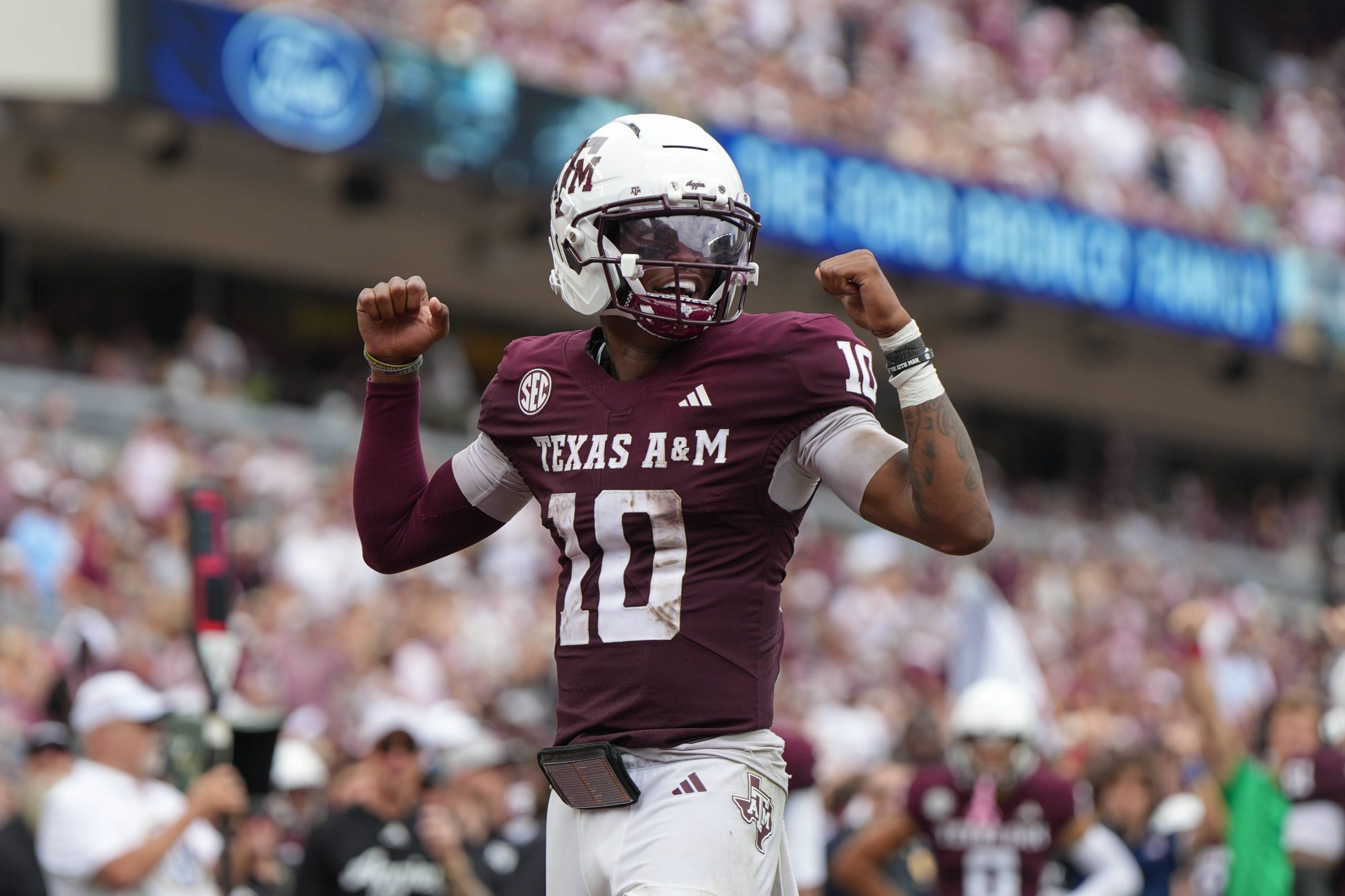 Ein Texas A&M-Football-Spieler, der eine kastanienbraune Uniform mit der Nummer 10, einen weißen Helm und ein Visier trägt, reckt während eines spannenden College Football Spiels in Week 2 jubelnd beide Arme über das Spielfeld. Der unscharfe Hintergrund zeigt eine Menge von Fans auf den Sitzen im Stadion. Diese Beschreibung wurde mit der FootballR KI automatisch generiert.