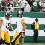 Zwei Footballspieler der Pittsburgh Steelers feiern während eines NFL-Spieltags auf dem Spielfeld. Beide tragen weiße Trikots mit gelben Hosen und schwarzen Helmen. Ein Spieler hält einen Football, während im Hintergrund Fans in grünen Trikots und Sicherheitskräfte zu sehen sind. Diese Beschreibung wurde mit der FootballR KI automatisch generiert.