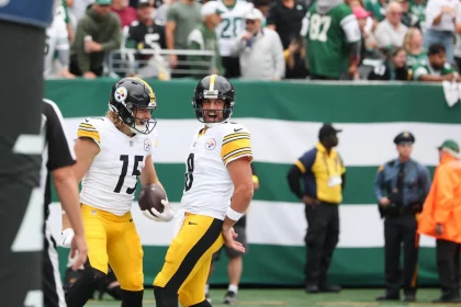 Zwei Footballspieler der Pittsburgh Steelers feiern während eines NFL-Spieltags auf dem Spielfeld. Beide tragen weiße Trikots mit gelben Hosen und schwarzen Helmen. Ein Spieler hält einen Football, während im Hintergrund Fans in grünen Trikots und Sicherheitskräfte zu sehen sind. Diese Beschreibung wurde mit der FootballR KI automatisch generiert.