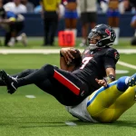 Ein Footballspieler in einem schwarzen Trikot der Houston Texans, C.J. Stroud, wird in der Luft von einem gelb-blauen Verteidiger der Los Angeles Rams auf einem grünen Feld angegriffen. Der Texans-Spieler hält den Ball fest umklammert, während er nach hinten fällt. Diese Beschreibung wurde mit der FootballR KI automatisch generiert.