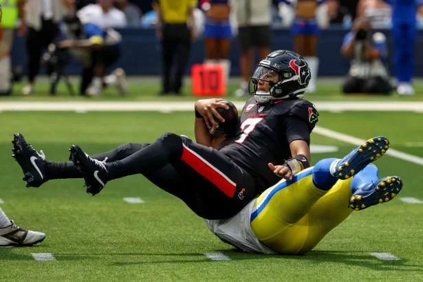 Ein Footballspieler in einem schwarzen Trikot der Houston Texans, C.J. Stroud, wird in der Luft von einem gelb-blauen Verteidiger der Los Angeles Rams auf einem grünen Feld angegriffen. Der Texans-Spieler hält den Ball fest umklammert, während er nach hinten fällt. Diese Beschreibung wurde mit der FootballR KI automatisch generiert.