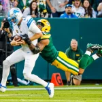 Micah Parsons in Grün und Gelb greift einen Spieler der Detroit Lions in Weiß an und schlingt seine Arme um die Taille des Gegners. Der Lions-Spieler, umklammert den Football fest, während beide fallen. Diese Beschreibung wurde mit der FootballR KI automatisch generiert.