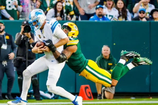 Micah Parsons in Grün und Gelb greift einen Spieler der Detroit Lions in Weiß an und schlingt seine Arme um die Taille des Gegners. Der Lions-Spieler, umklammert den Football fest, während beide fallen. Diese Beschreibung wurde mit der FootballR KI automatisch generiert.
