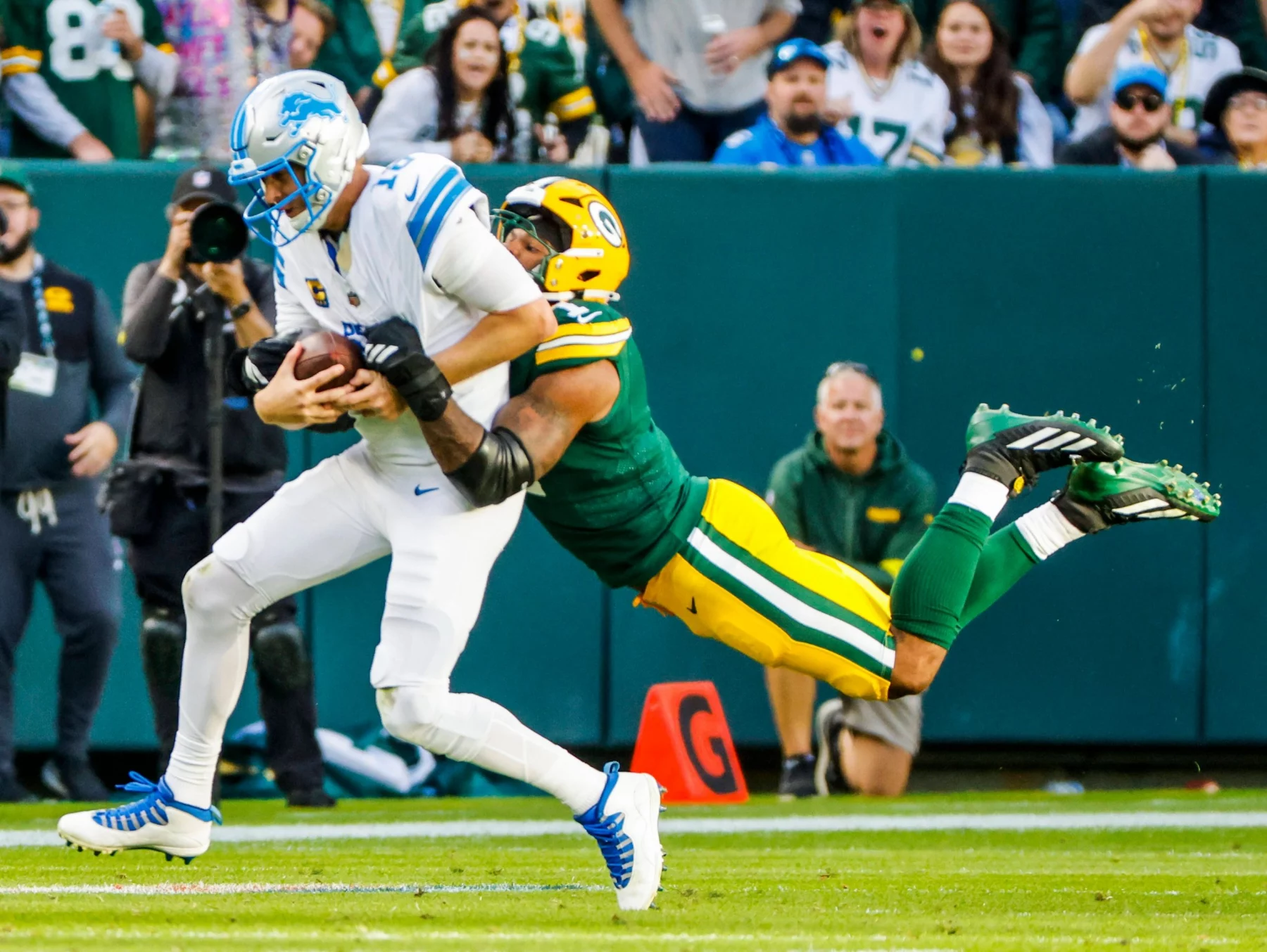 Micah Parsons in Grün und Gelb greift einen Spieler der Detroit Lions in Weiß an und schlingt seine Arme um die Taille des Gegners. Der Lions-Spieler, umklammert den Football fest, während beide fallen. Diese Beschreibung wurde mit der FootballR KI automatisch generiert.