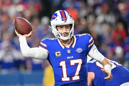 Josh Allen, der seine blaue Bills-Uniform mit der Nummer 17 trägt, bereitet sich darauf vor, während eines Spiels einen Pass zu werfen. Er trägt einen weißen Helm mit dem rot-blauen Buffalo-Logo, während im Hintergrund unscharfe Teamkollegen und das Stadionpublikum zu sehen sind. Diese Beschreibung wurde mit der FootballR KI automatisch generiert.