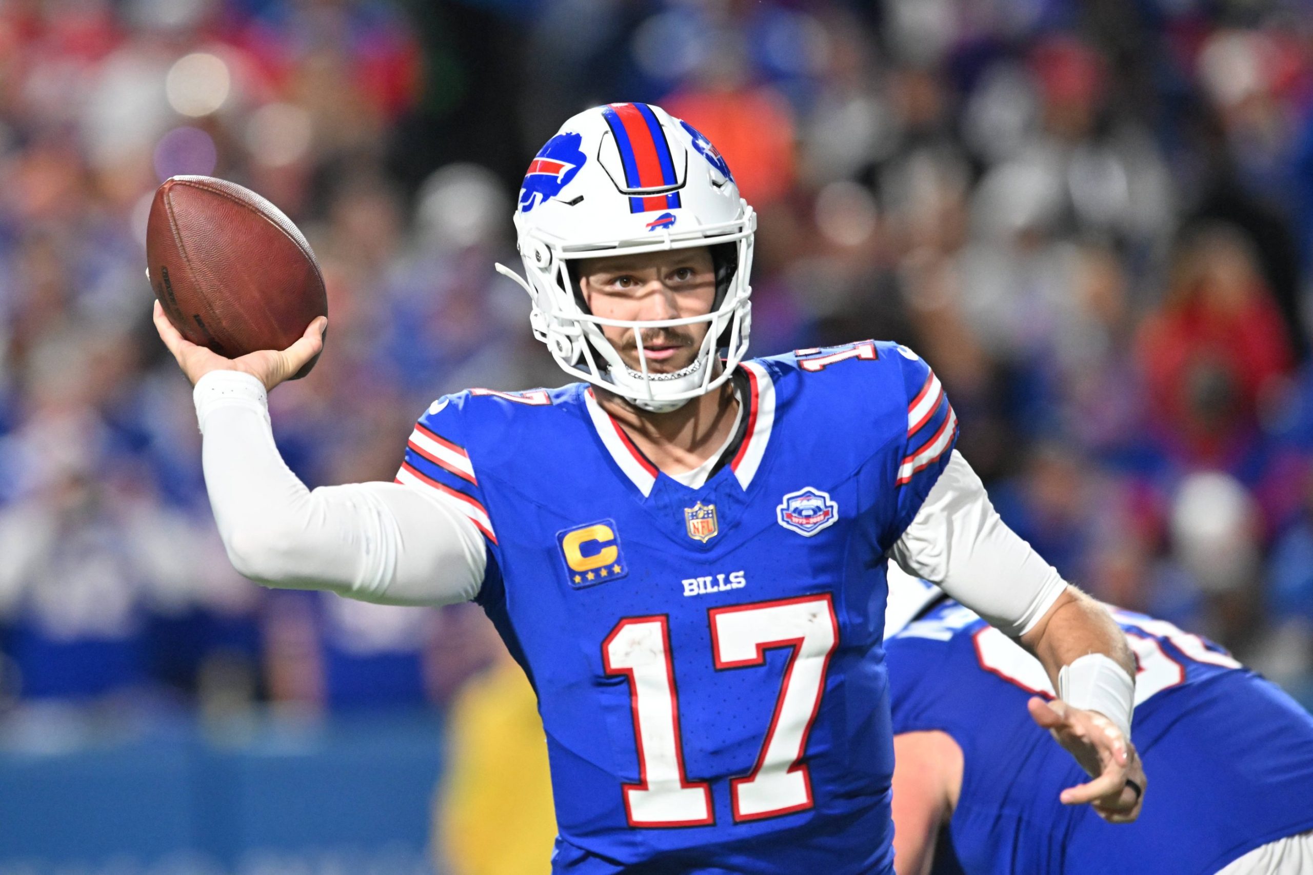 Josh Allen, der seine blaue Bills-Uniform mit der Nummer 17 trägt, bereitet sich darauf vor, während eines Spiels einen Pass zu werfen. Er trägt einen weißen Helm mit dem rot-blauen Buffalo-Logo, während im Hintergrund unscharfe Teamkollegen und das Stadionpublikum zu sehen sind. Diese Beschreibung wurde mit der FootballR KI automatisch generiert.
