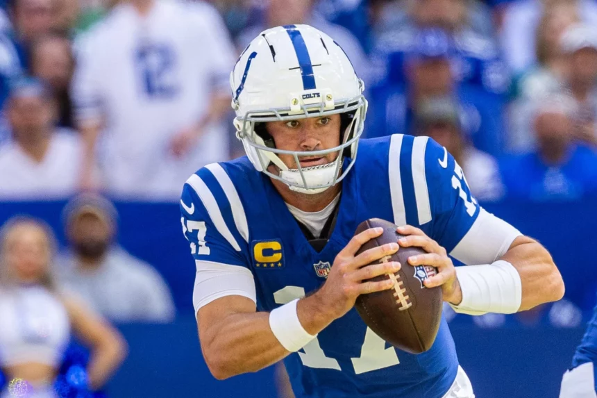 Daniel Jones in einer blauen Uniform der Indianapolis Colts und mit weißem Helm bereitet sich darauf vor, während der NFL Week 1 einen Football zu werfen. Er ist konzentriert und hält den Ball mit beiden Händen, die Nummer 17 ist zwischen Fans und Teamkameraden im Stadionhintergrund zu sehen. Diese Beschreibung wurde mit der FootballR KI automatisch generiert.