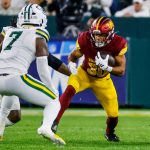 Austin Ekeler - Ein Footballspieler der Washington Commanders in einem rot-gelben Trikot rennt mit dem Ball, verfolgt von einem Verteidiger der Green Bay Packers, die beide trotz des allgegenwärtigen Risikos von Verletzungen wie einem Achillessehnenriss auf einem grünen Footballfeld Höchstleistungen erbringen. Diese Beschreibung wurde mit der FootballR KI automatisch generiert.
