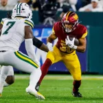 Austin Ekeler - Ein Footballspieler der Washington Commanders in einem rot-gelben Trikot rennt mit dem Ball, verfolgt von einem Verteidiger der Green Bay Packers, die beide trotz des allgegenwärtigen Risikos von Verletzungen wie einem Achillessehnenriss auf einem grünen Footballfeld Höchstleistungen erbringen. Diese Beschreibung wurde mit der FootballR KI automatisch generiert.