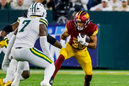 Austin Ekeler - Ein Footballspieler der Washington Commanders in einem rot-gelben Trikot rennt mit dem Ball, verfolgt von einem Verteidiger der Green Bay Packers, die beide trotz des allgegenwärtigen Risikos von Verletzungen wie einem Achillessehnenriss auf einem grünen Footballfeld Höchstleistungen erbringen. Diese Beschreibung wurde mit der FootballR KI automatisch generiert.