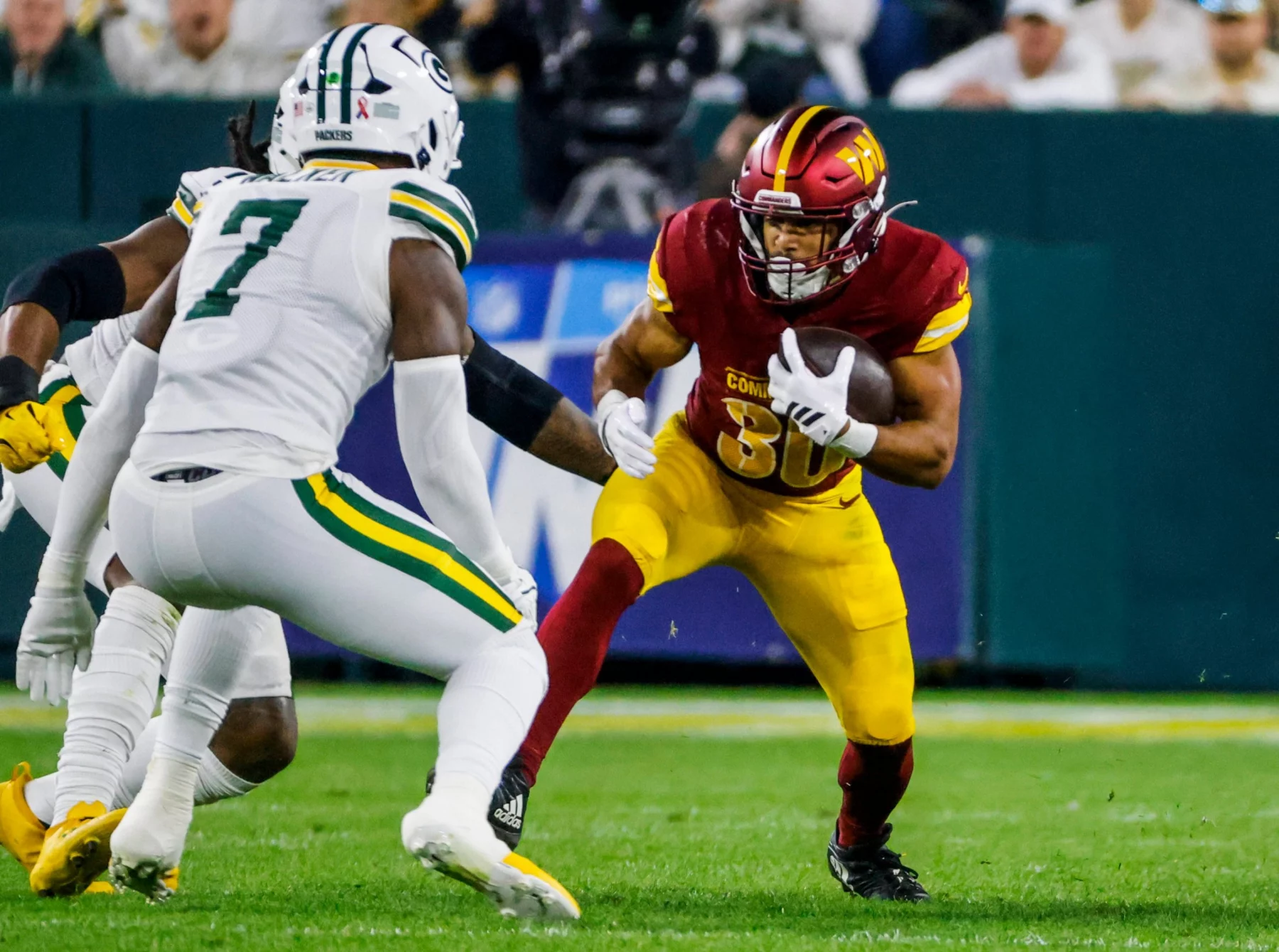 Austin Ekeler - Ein Footballspieler der Washington Commanders in einem rot-gelben Trikot rennt mit dem Ball, verfolgt von einem Verteidiger der Green Bay Packers, die beide trotz des allgegenwärtigen Risikos von Verletzungen wie einem Achillessehnenriss auf einem grünen Footballfeld Höchstleistungen erbringen. Diese Beschreibung wurde mit der FootballR KI automatisch generiert.