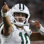 Jordan Love, der ein weißes Green Bay Packers-Trikot und einen Helm mit der Nummer 10 trägt, jubelt auf dem Spielfeld und zeigt mit beiden Händen nach vorne. Der Hintergrund ist unscharf mit Stadionlichtern und Fans. Diese Beschreibung wurde mit der FootballR KI automatisch generiert.