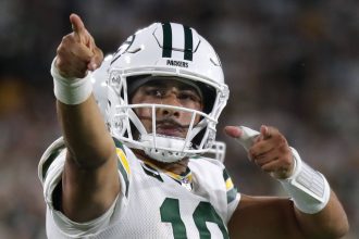 Bye Week - Jordan Love, der ein weißes Green Bay Packers-Trikot und einen Helm mit der Nummer 10 trägt, jubelt auf dem Spielfeld und zeigt mit beiden Händen nach vorne. Der Hintergrund ist unscharf mit Stadionlichtern und Fans. Diese Beschreibung wurde mit der FootballR KI automatisch generiert.
