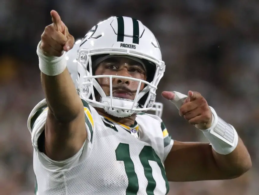 Jordan Love, der ein weißes Green Bay Packers-Trikot und einen Helm mit der Nummer 10 trägt, jubelt auf dem Spielfeld und zeigt mit beiden Händen nach vorne. Der Hintergrund ist unscharf mit Stadionlichtern und Fans. Diese Beschreibung wurde mit der FootballR KI automatisch generiert.