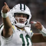 Langer Weg nach Phoenix - Bye Week - Jordan Love, der ein weißes Green Bay Packers-Trikot und einen Helm mit der Nummer 10 trägt, jubelt auf dem Spielfeld und zeigt mit beiden Händen nach vorne. Der Hintergrund ist unscharf mit Stadionlichtern und Fans. Diese Beschreibung wurde mit der FootballR KI automatisch generiert.