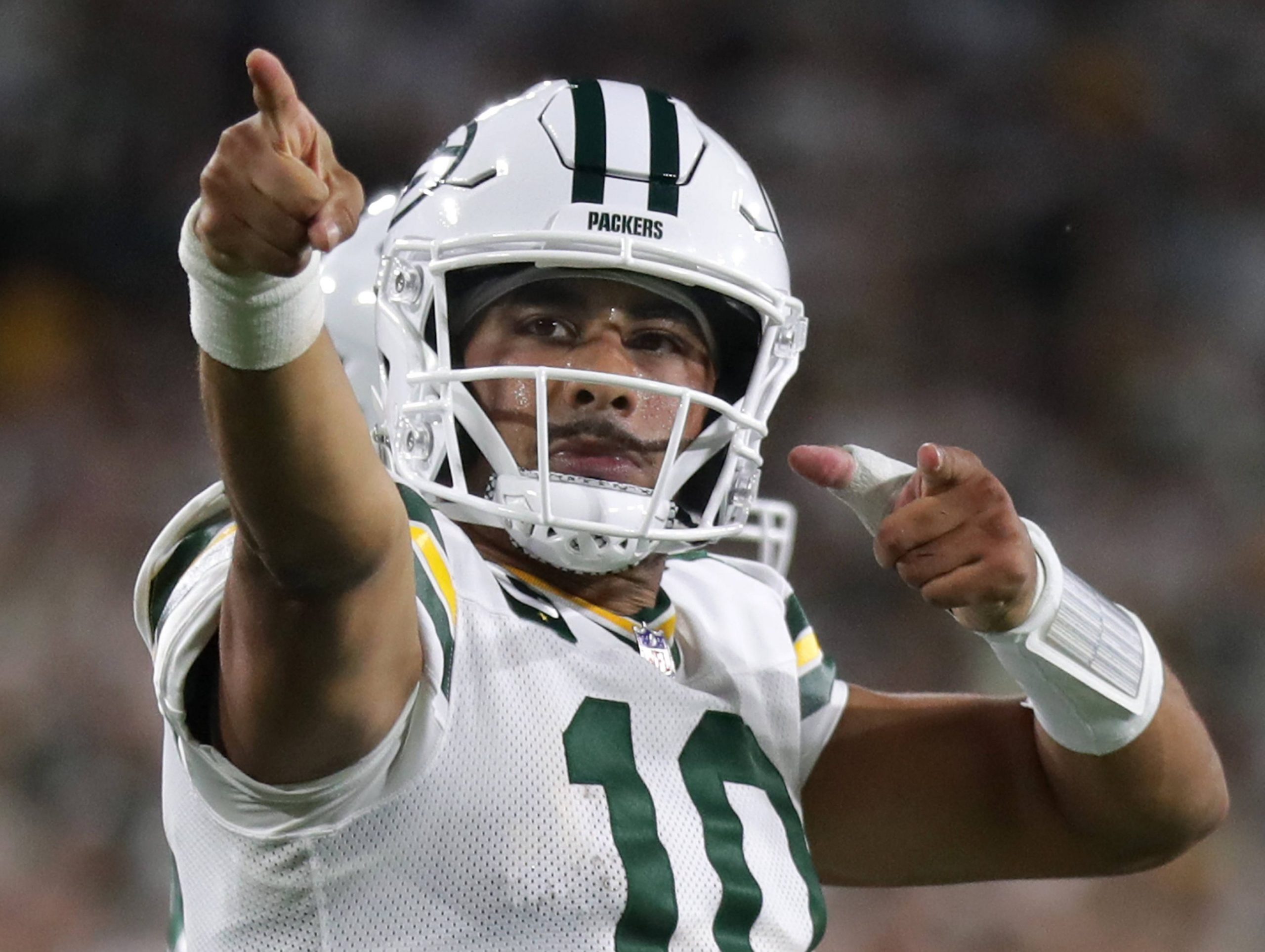 Bye Week - Jordan Love, der ein weißes Green Bay Packers-Trikot und einen Helm mit der Nummer 10 trägt, jubelt auf dem Spielfeld und zeigt mit beiden Händen nach vorne. Der Hintergrund ist unscharf mit Stadionlichtern und Fans. Diese Beschreibung wurde mit der FootballR KI automatisch generiert.