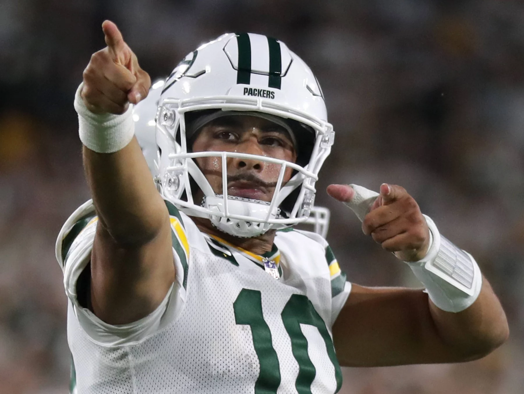 Langer Weg nach Phoenix - Bye Week - Jordan Love, der ein weißes Green Bay Packers-Trikot und einen Helm mit der Nummer 10 trägt, jubelt auf dem Spielfeld und zeigt mit beiden Händen nach vorne. Der Hintergrund ist unscharf mit Stadionlichtern und Fans. Diese Beschreibung wurde mit der FootballR KI automatisch generiert.