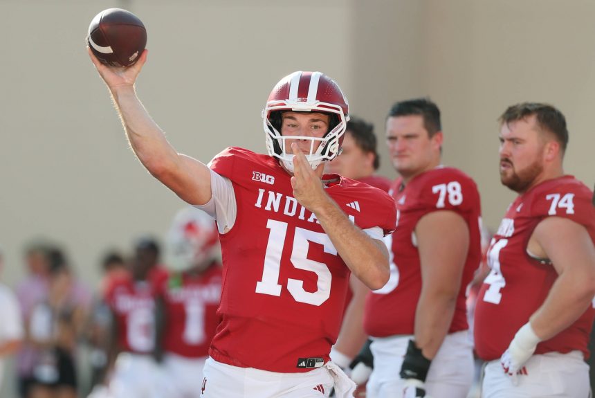 College Football Woche 3 -Ein Football-Spieler in einem roten Indiana-Trikot mit der Nummer 15 bereitet sich während der College Football Woche 3 auf den Wurf vor. Er trägt einen Helm, während seine Teamkollegen in roten Trikots mit den Nummern 78 und 74 hinter ihm stehen. Andere Spieler sind im Hintergrund zu sehen. Diese Beschreibung wurde mit der FootballR KI automatisch generiert.