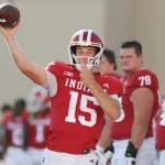 College Football Woche 3 -Ein Football-Spieler in einem roten Indiana-Trikot mit der Nummer 15 bereitet sich während der College Football Woche 3 auf den Wurf vor. Er trägt einen Helm, während seine Teamkollegen in roten Trikots mit den Nummern 78 und 74 hinter ihm stehen. Andere Spieler sind im Hintergrund zu sehen. Diese Beschreibung wurde mit der FootballR KI automatisch generiert.