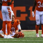 NFL Spieltag 2 - Joe Burrow im Bengals-Trikot mit der Nummer 9 sitzt nach dem NFL-Spieltag 2 auf dem Spielfeld und sieht verzweifelt aus. Sein Helm liegt auf dem Boden, während zwei orange-weiß gekleidete Mannschaftskameraden die Sicht teilweise versperren, wobei der Rasen und ein großes oranges Schild zu sehen sind. Diese Beschreibung wurde mit der FootballR KI automatisch generiert.