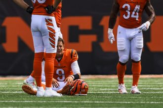 NFL Spieltag 2 - Joe Burrow im Bengals-Trikot mit der Nummer 9 sitzt nach dem NFL-Spieltag 2 auf dem Spielfeld und sieht verzweifelt aus. Sein Helm liegt auf dem Boden, während zwei orange-weiß gekleidete Mannschaftskameraden die Sicht teilweise versperren, wobei der Rasen und ein großes oranges Schild zu sehen sind. Diese Beschreibung wurde mit der FootballR KI automatisch generiert.